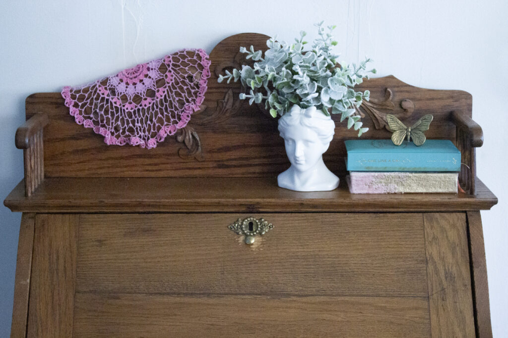 Vintage secretary desk styled with a bust, books, a butterfly, and a pink doily